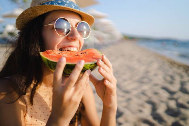 Young, happy, woman eating watermelon on the beach, wearing sunglasses.