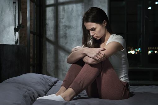 Young female sitting on bed holding knees thinking about some problems with miserable face